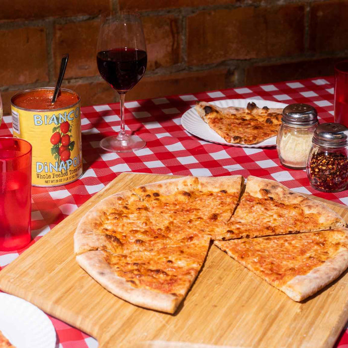 New York-style cheese pizza on an Ooni Bamboo  Pizza Peel & Serving Board on a table next to slices of pizza, a can of Bianco di Napoli tomatoes and glasses.
