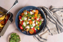 Slow-roasted squash, eggplant, onions, cherry tomatoes and sweet peppers in an Ooni Roasting pan next to a bowl of slow-roasted vegetables with feta and fresh herbs on a table.