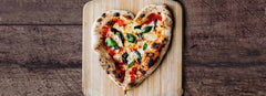 A heart shaped pizza topped with tomato, cheese and basil on a wooden pizza peel