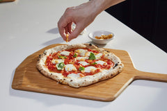 Pizza topped with cheese, tomatoes, basil being sprinkled with fried garlic chips on a wooden pizza peel