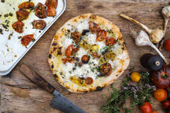 Cooked pizza with slow-roasted tomatoes and garlic on a table next to an assortment of tomatoes, garlic bulbs, a knife, and a baking tray with roasted tomatoes.