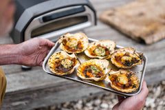 Two hands holding a roasting tray with roasted hand-dived scallops with chorizo, garlic and parsley.