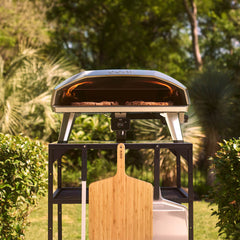 An outdoor setup featuring an Ooni Koda 2 Max pizza oven on a black stand. The oven top shows the 'ooni' logo, and a digital display reads '458'. A wooden pizza peel with the 'ooni' logo is positioned in the foreground.