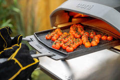 Two hands wearing Ooni Pizza Oven Gloves pulling out a Dual-Sided Grizzler Plate with roasted tomatoes from an Ooni Koda 16.