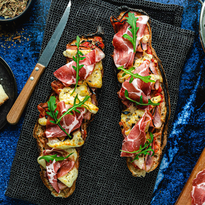 Cooked Corsican pizza baguette with Corsican cheese, pancetta, coppa, arugula and herbes de Provence on a table surrounded by a plate of cheese, bowl of arugula and serving board of pancetta.