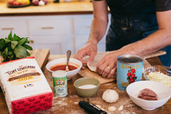 Several different types of pizza ingredients, like flour, tomatoes and yeast on a wooden worktop with a man stretching pizza dough