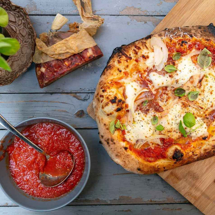 Pizza topped with tomatoes, cheese and soppressata on a wooden chopping board next to a bowl of tomato pizza sauce. Baked using a soppressata pizza recipe.