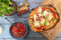 Pizza topped with tomatoes, cheese and soppressata on a wooden chopping board next to a bowl of tomato pizza sauce. Baked using a soppressata pizza recipe.