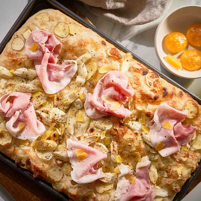 Pizza bianca with white asparagus, sliced potatoes, ham and cured egg next to a bowl with cured egg yolks.