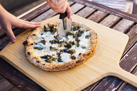 A hand cutting a cooked calzone with escarole and provola on top of an Ooni Bamboo Pizza Peel on a table.