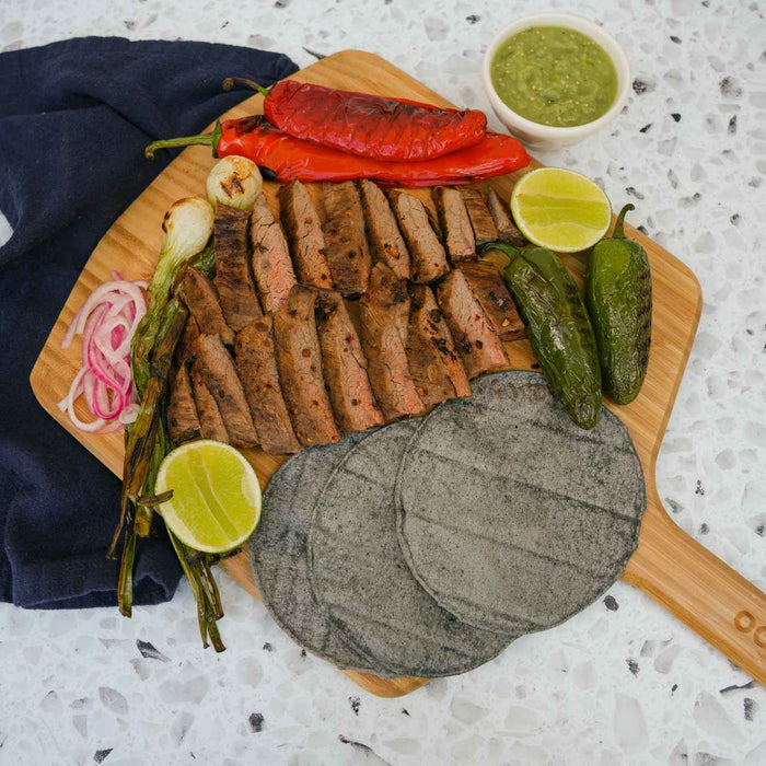 Masienda blue corn tortillas on an Ooni Bamboo Pizza Peel & Serving Board next to green and red chilies, sliced carne asada, grilled onions, sliced limes and salsa verde.