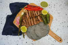 Masienda blue corn tortillas on an Ooni Bamboo Pizza Peel & Serving Board next to green and red chilies, sliced carne asada, grilled onions, sliced limes and salsa verde.