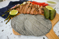 Masienda blue corn tortillas on an Ooni Bamboo Pizza Peel & Serving Board next to green and red chilies, sliced carne asada, grilled onions, sliced limes and salsa verde.