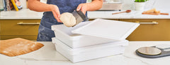 person lifting dough ball from ooni dough boxes in kitchen setting with floured bamboo pizza peel on marble counter