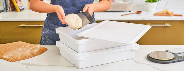 person lifting dough ball from ooni dough boxes in kitchen setting with floured bamboo pizza peel on marble counter
