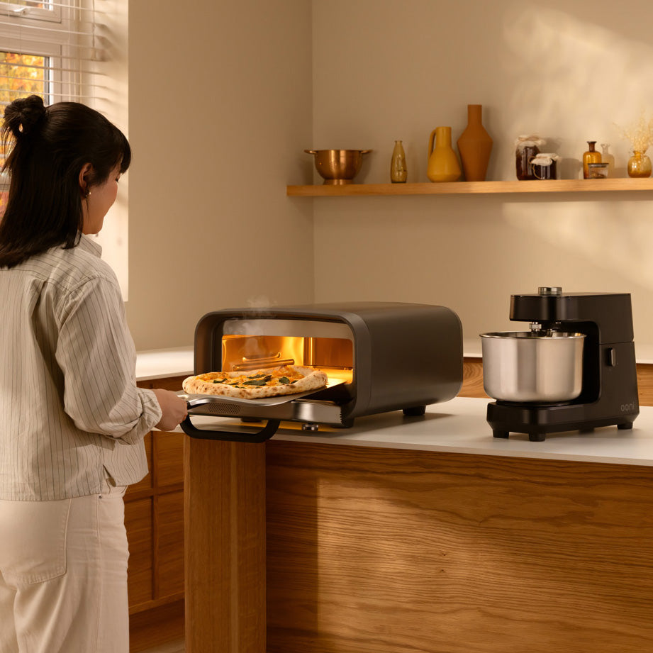 A woman in a kitchen pulls a pizza out of a ooni volt 2 electric pizza oven with a pizza peel. A black ooni halo core stand mixer with a stainless steel bowl sits on the counter beside it.