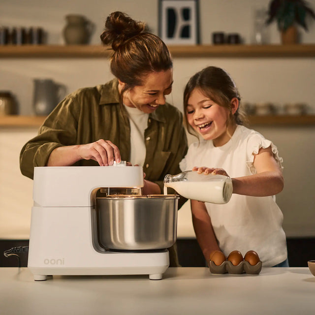 A woman and a young girl are baking together in a warm kitchen setting. The woman is wearing an olive green shirt, and the girl is in a white ruffled top.