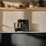 Ooni halo core spiral mixer with a stainless steel bowl sits on a kitchen counter, with warm sunlight casting windowpane shadows on the background wall and wooden shelf.