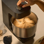 A black stand mixer with a stainless steel bowl containing dough, with a hand adjusting the top dial. A small bowl and a black spatula are visible on a light beige surface.