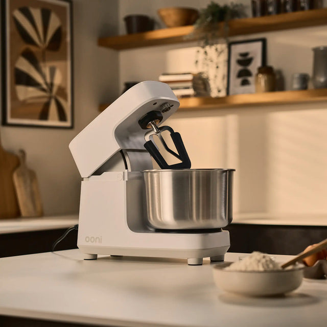 Ooni halo core white stand mixer with a stainless steel bowl sits on a kitchen counter, with a bowl of flour in the foreground and a softly blurred kitchen interior behind it.