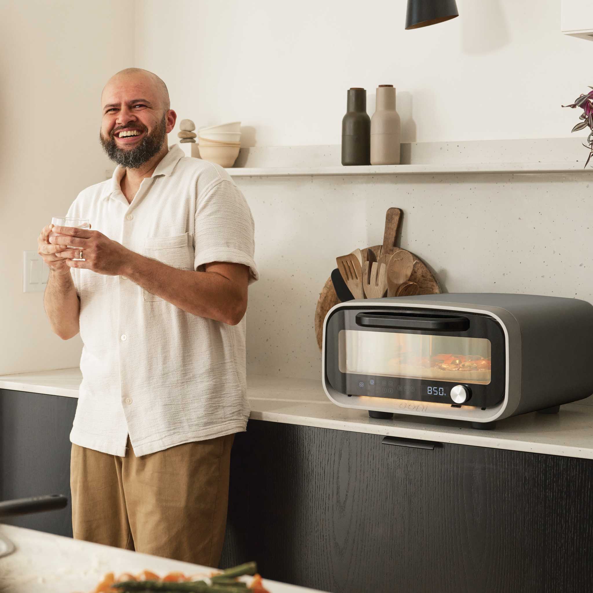 Man enjoying a glass of wine beside an Ooni Volt 2 electric pizza oven on a kitchen countertop.