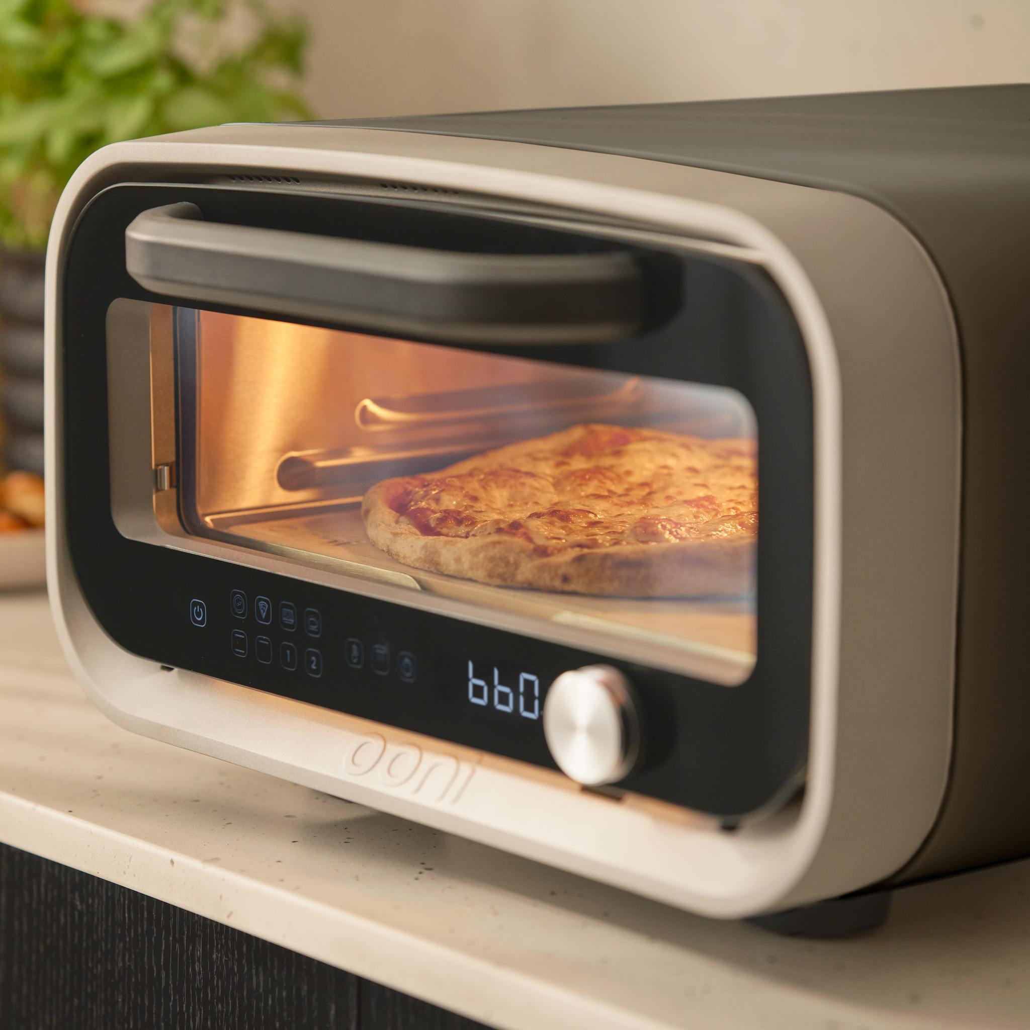 Cheese pizza baking inside the Ooni Volt 2 electric pizza oven, visible through the glass window.