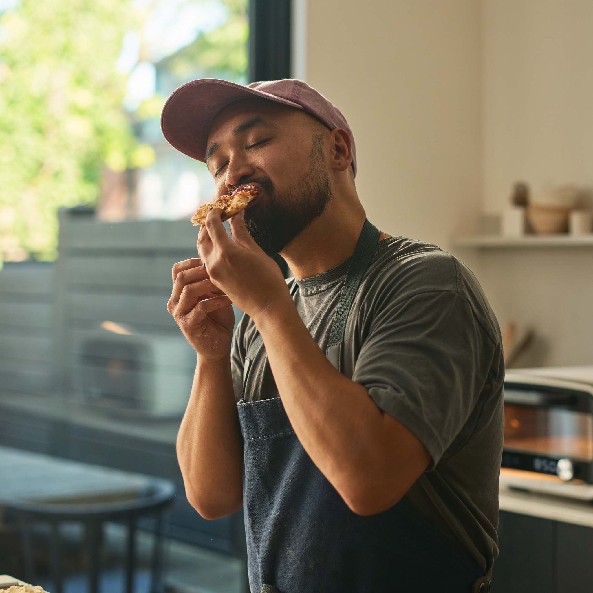 Man eating slice of pizza with eyes closed inside modern kitchen. 