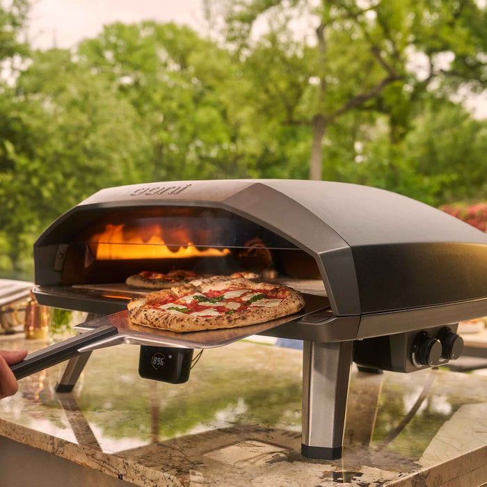 Ooni Koda 2 Max outdoor pizza oven cooking two pizzas, one already in the oven and another being inserted on a pizza peel in a backyard. | Click this image to open up the product gallery modal. The product gallery modal allows the images to be zoomed in on.